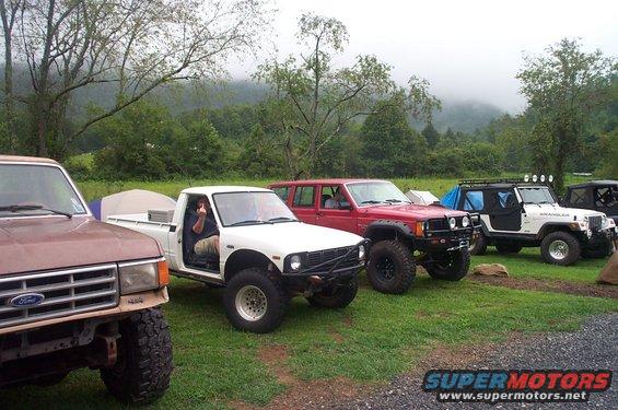 Several off-road vehicles, including a brown Ford truck, a white pickup, a red SUV, and a white Jeep Wrangler, parked on a grassy area with trees and hills in the background