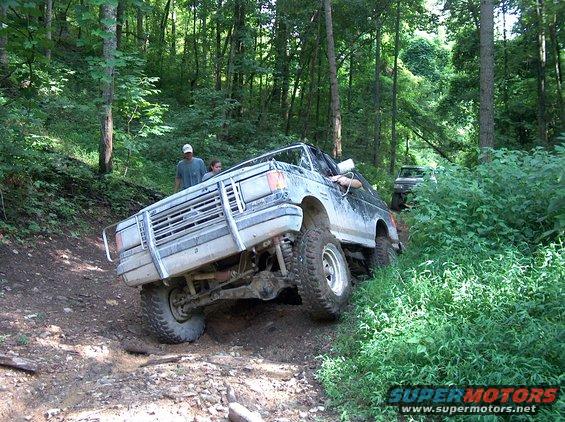 1989 Ford Bronco tilted on a muddy trail with another vehicle behind it in the woods