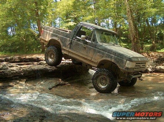 Mud-covered Ford Bronco navigating a rocky stream, front-right tire elevated