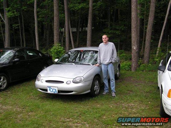 Man standing by silver 1998 Ford Taurus, parked on grass with trees behind. Black car to left, white car to right