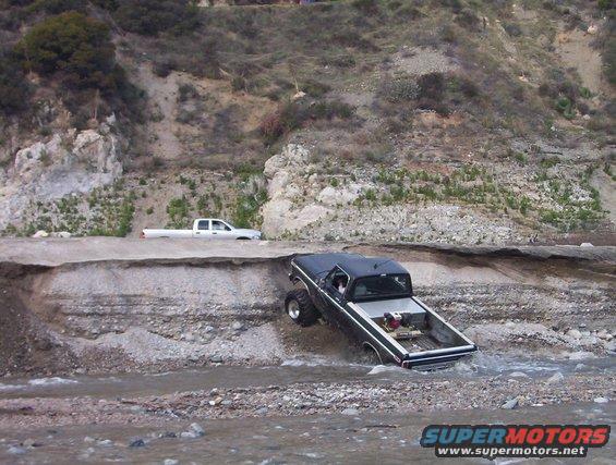 Black pickup truck with large tires driving through water, a white pickup truck is on a ledge above