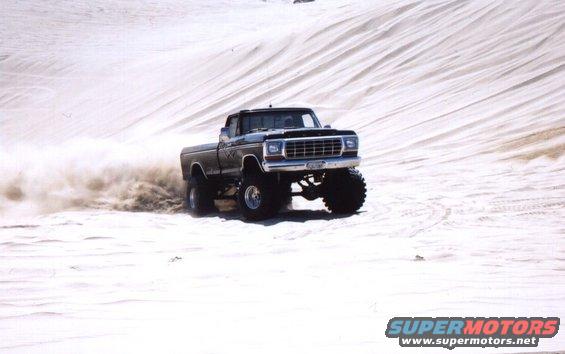 Lifted pickup truck kicking up dust while driving on sand dunes. Appears to be a 1979 Ford F-Series
