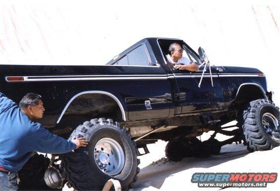 Black pickup truck with large off-road tires, lifted suspension, and chrome trim. A man is in the driver's seat, and another is near the front wheel