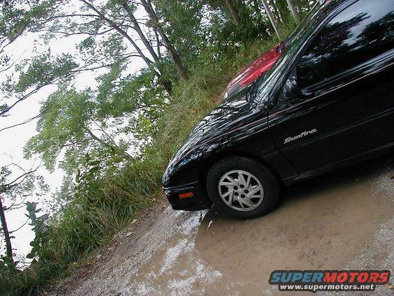 Black Pontiac Sunfire with red car behind it, parked on a muddy road with trees and water