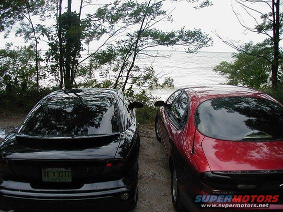 Two sedans, one black and one red, parked on a dirt road next to a large body of water with trees in the background