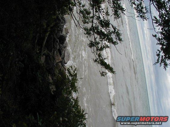 Shoreline with choppy water, rocks, and trees under a cloudy sky. Supermotors.net logo in lower right
