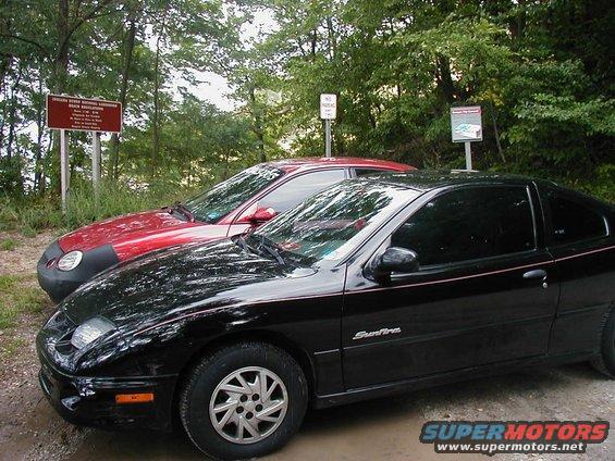 Black Pontiac Sunfire coupe and red car parked on dirt road with trees and signs