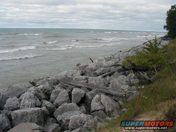 Rocky shoreline with driftwood and choppy water under a cloudy sky. Website watermark at bottom right