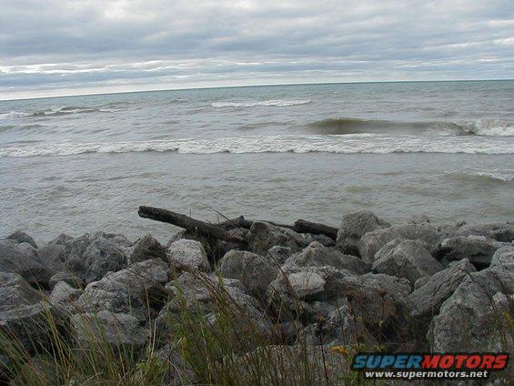Rocky shoreline with waves under a cloudy sky. Watermark reads SUPERMOTORS