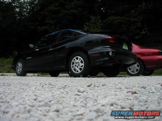 Black car parked next to a red car on a gravel surface with trees in the background