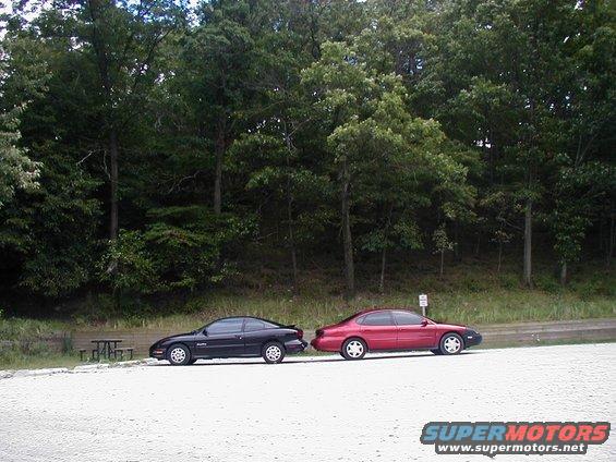 Red 1997 Ford Taurus parked on a gravel lot next to a black coupe