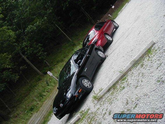 Black car and red car parked on a gravel lot next to trees