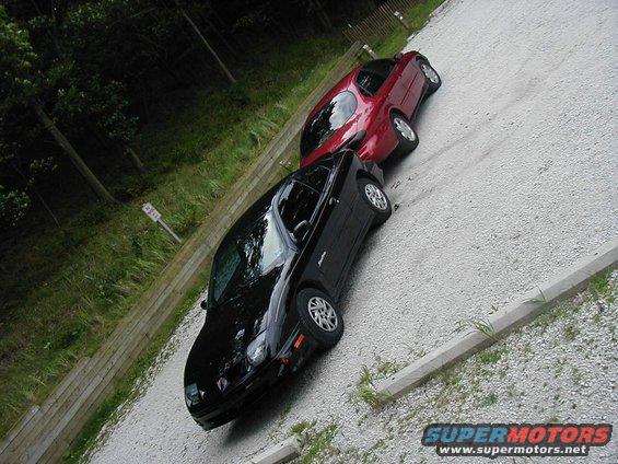 Black and red cars parked on a gravel lot next to a wooden fence and trees