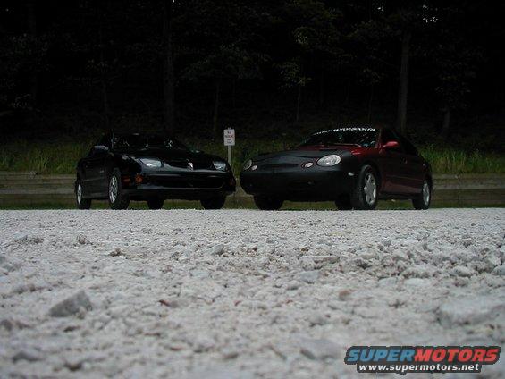 Two cars, a black Pontiac Firebird and a red car with a black cover, parked side-by-side on a gravel lot