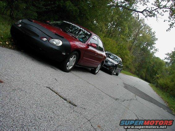 Red 1997 Ford Taurus with car bra, parked on a road with another car behind it