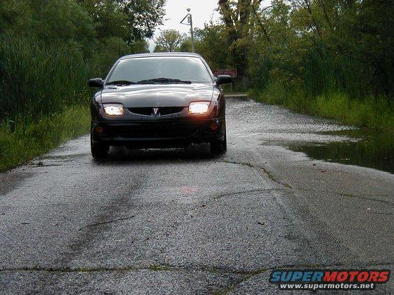 Black Pontiac Sunfire driving on a wet road with trees on either side