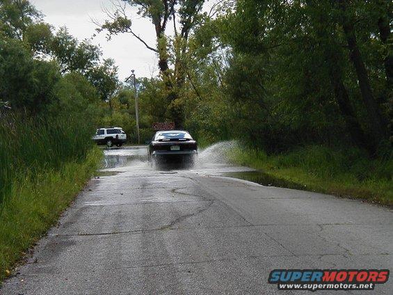 Dark sedan driving through water, kicking up spray. White SUV visible in background