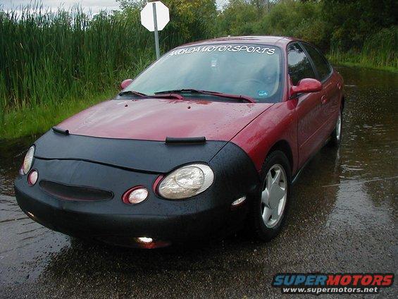Red sedan with a black car bra on the front, parked in a wet area. Windshield banner says 'FLOWDA MOTORSPORTS'