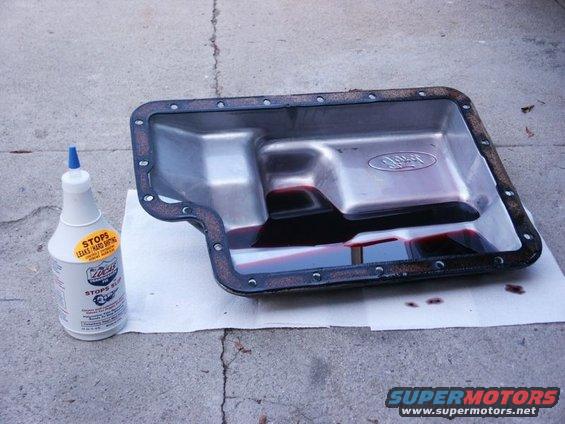 Metal transmission pan with dark red fluid and a bottle of Lucas Stop Leak on white paper