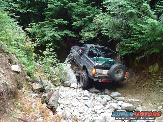 1993 Ford Bronco navigating a steep, rocky off-road trail with dense forest on either side