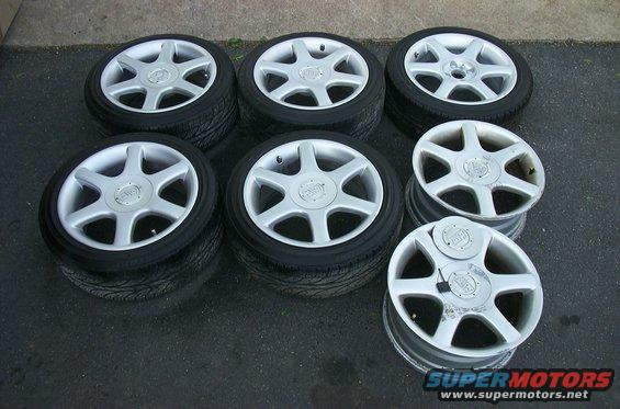 Seven silver alloy wheels with tires stacked on asphalt. Some wheels show scuff marks