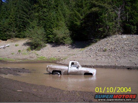 White pickup truck stuck in muddy water with trees and a rocky hill in the background