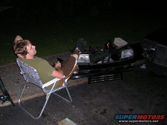 Person relaxing in a lawn chair beside the front end of a car with its hood open
