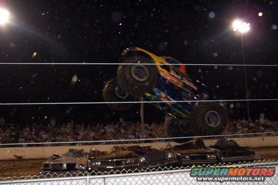 Monster truck airborne over a pile of crushed cars at night, crowd in background