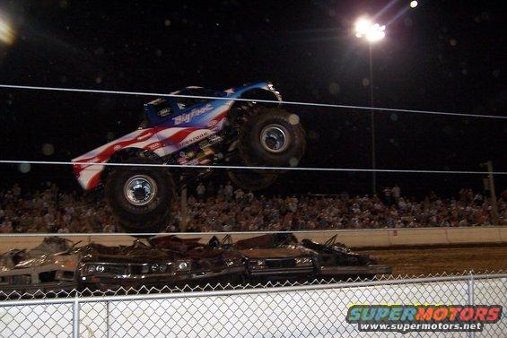 Monster truck with American flag paint scheme, labeled Bigfoot, airborne over a pile of wrecked cars in front of a crowd