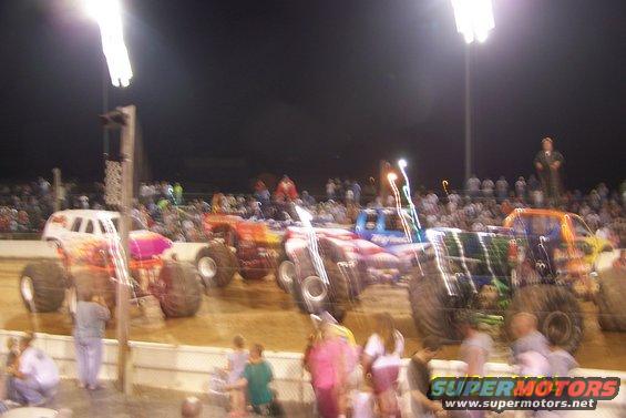 Multiple monster trucks lined up on a dirt track with crowds watching under stadium lights