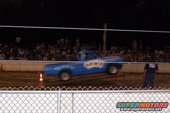 Blue pickup truck with 'JOKER' text and playing card graphics on its side, driving on a ramp at night