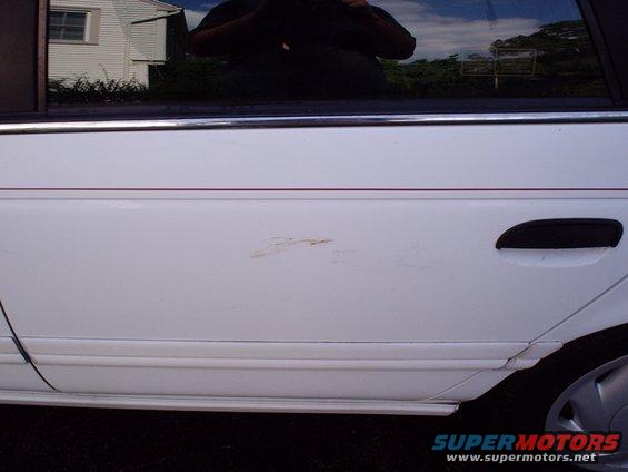 White car door showing a rust spot and a black door handle. A thin red pinstripe runs horizontally above the rust