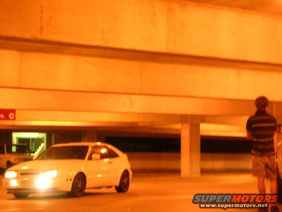 White coupe with headlights on in a parking garage. Another car is visible in the background