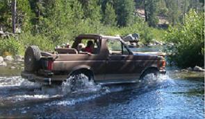 Brown 1990 Ford Bronco driving through a shallow river with trees in the background
