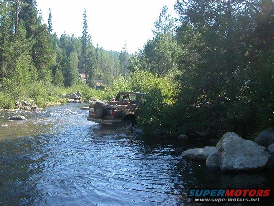 Brown Ford Bronco driving through a shallow river, spare tire mounted on the rear door