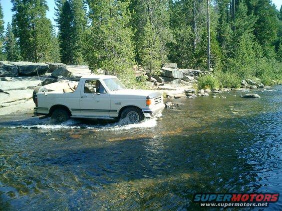 1990 Ford Bronco driving through a shallow river with trees and rocks in the background