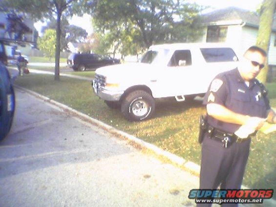 White Ford Bronco parked on grass, front 3/4 view. Police officer stands nearby