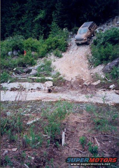 Brown Ford Bronco climbing a steep, rocky, dirt hill with trees in the background