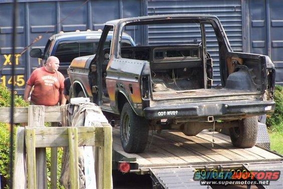 Rear view of a stripped-down 1978 Ford Bronco on a trailer, with a man standing nearby and another truck behind it