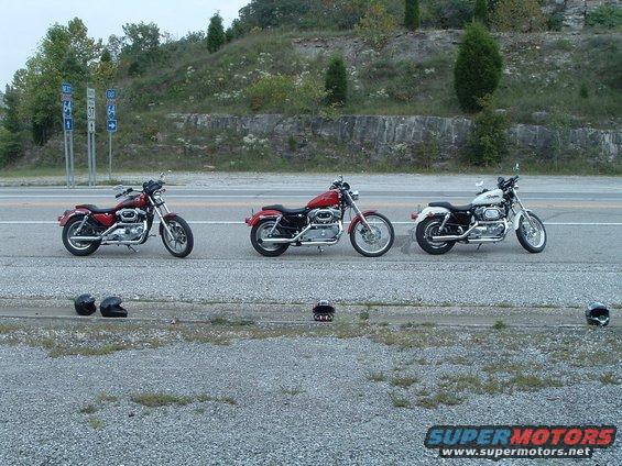 Three motorcycles parked on the side of a road. Two red, one white. Helmets are on the ground