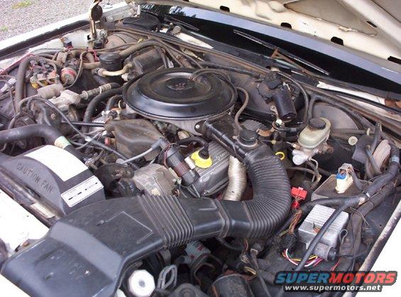 Engine bay of a car with various hoses, wires, and engine components visible. Includes a black air filter housing and a radiator fan