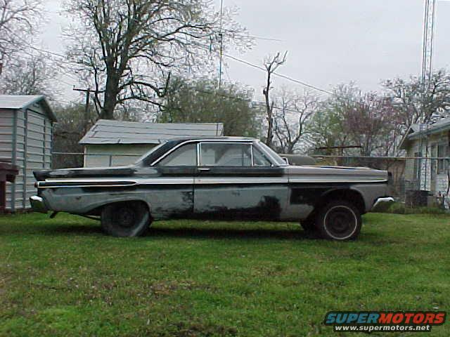 side view of a beat-up 1960s sedan with primer spots and flat tires, parked on grass near sheds