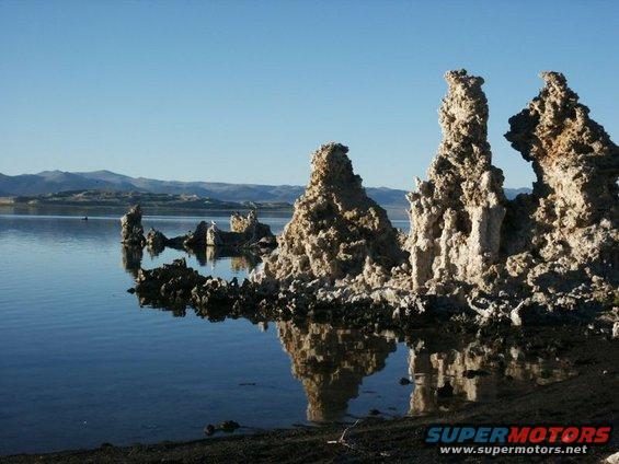 Tufa formations rise from a calm lake under a clear blue sky. Mountains are visible in the distance