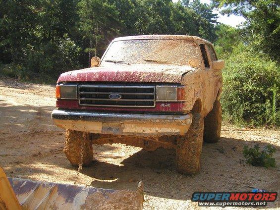 Mud-covered red 1987 Ford Bronco front view, parked on dirt with trees in background
