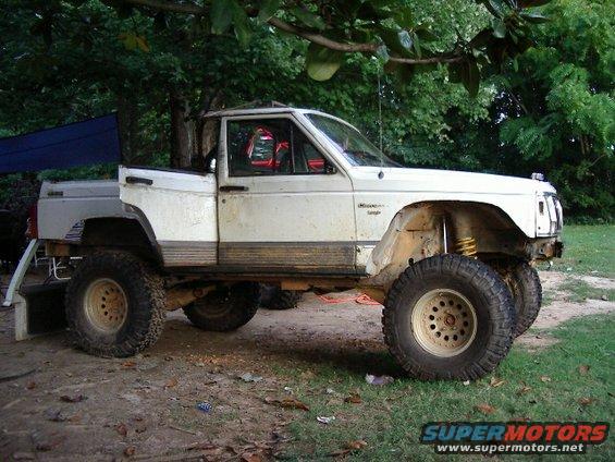 White Jeep Comanche pickup truck with large off-road tires, lifted suspension, and some body damage. It's parked outdoors under trees