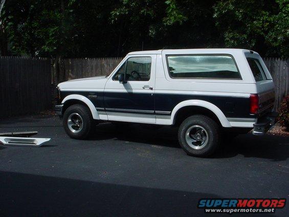 Two-tone white and dark blue Ford Bronco parked on asphalt, with a wooden fence and trees in the background