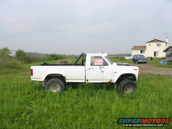White pickup truck with off-road tires and roll bar, parked in grass. Another dark SUV is visible in the background near a house