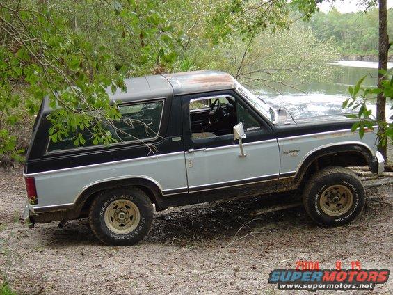 Two-tone gray and black Ford Bronco with a removable hardtop, parked on a dirt path near water
