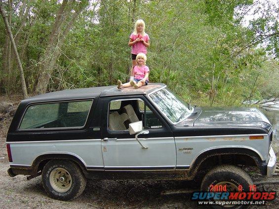 Two-tone gray and black Ford Bronco, side view, with two girls on the roof