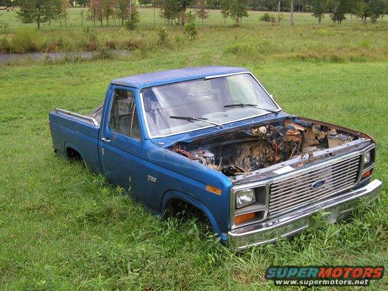 Blue pickup truck with hood open, sitting in tall grass. Engine bay is empty